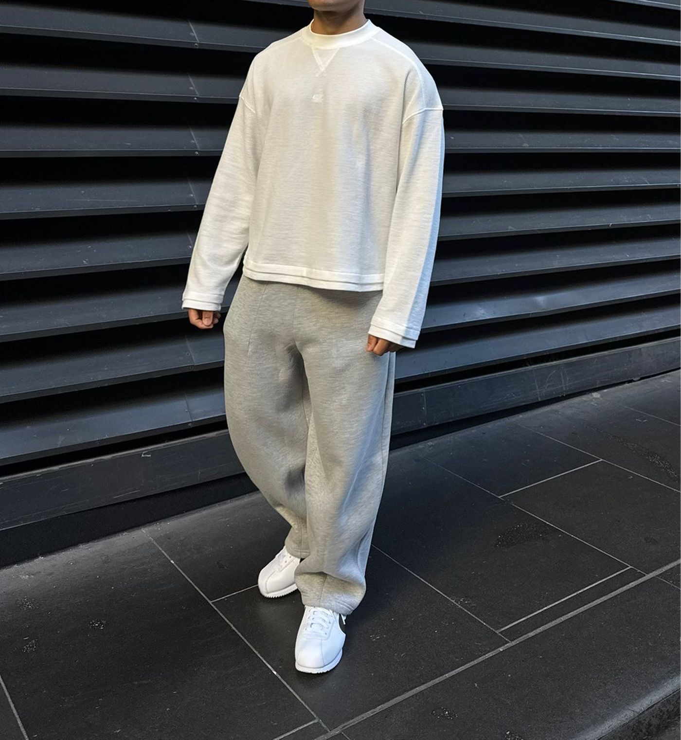 A person wearing a light off-white long-sleeve cropped sweatshirt and light gray wide-leg sweatpants, standing against a dark gray slatted wall.